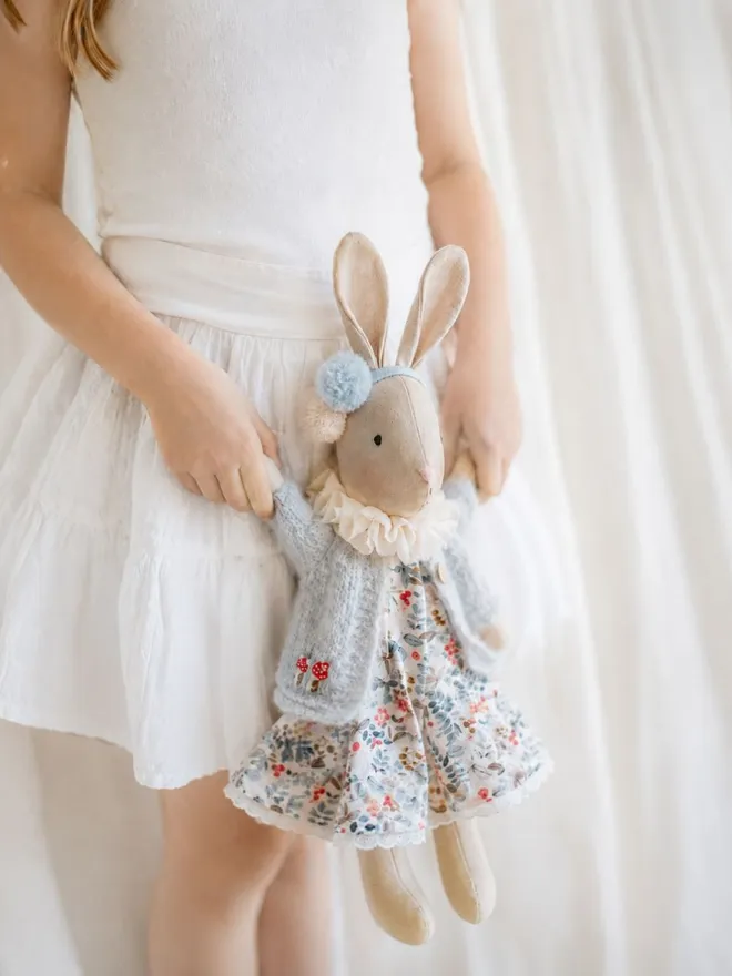Young girl in a white dress holding a handmade Verity Isabelle Veronica Bunny wearing a blue floral dress, soft knitted cardigan and pom-pom headband.