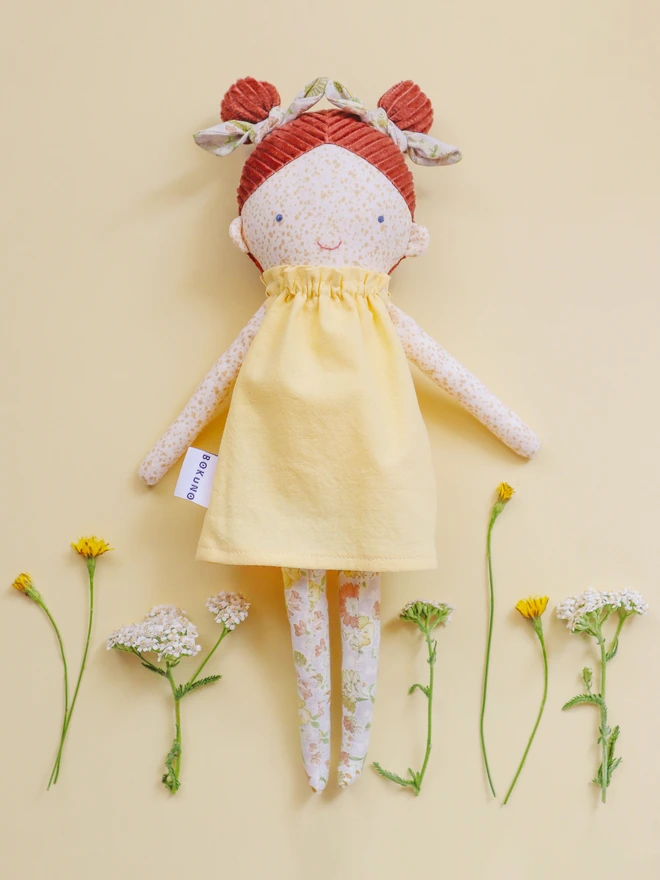 Handmade heirloom cloth doll with freckled face, red corduroy hair in two buns, floral leggings, and a soft yellow dress, styled on a neutral background with delicate wildflowers.