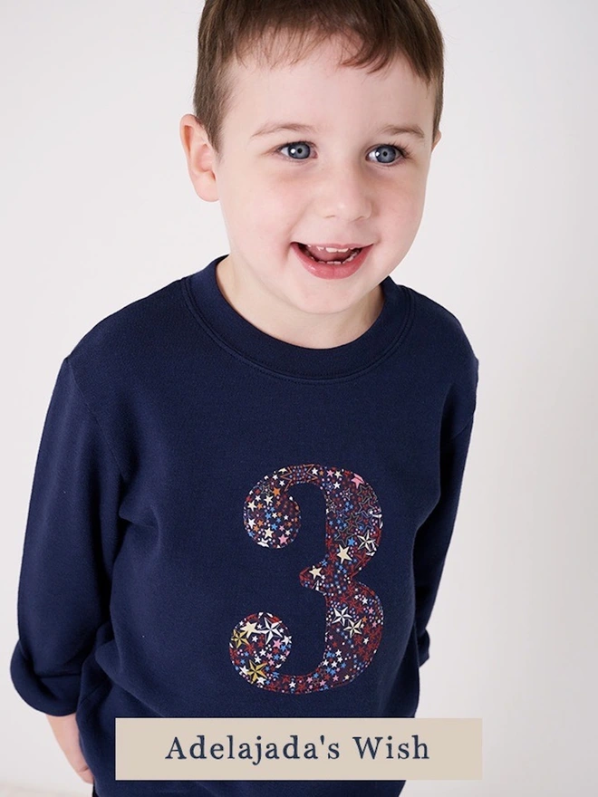  A 3 year old boy wearing a navy birthday t-shirt with a number 3 cut from liberty starl print on the front