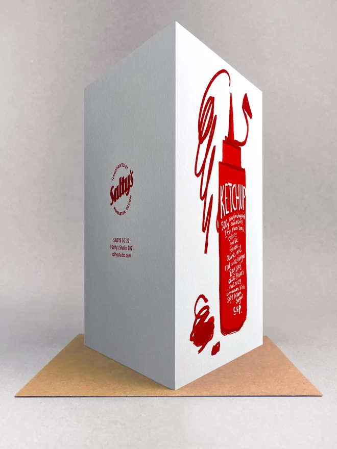 Rear view of Ketchup recipe card, written on a squeezy red bottle, with a scribbled squirt of ketchup jumping out the top - all on a white greetings card stood on a brown kraft envelope. All in a light grey studio space.