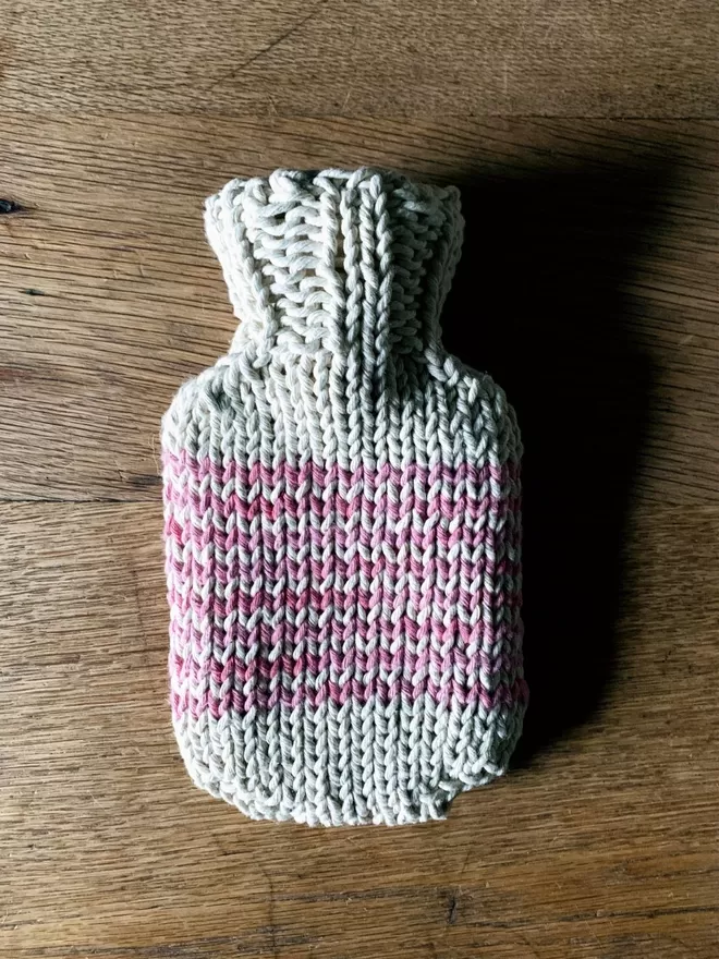 Mini Hot Water Bottle in hand knit string cover with pink stripe on an oak table