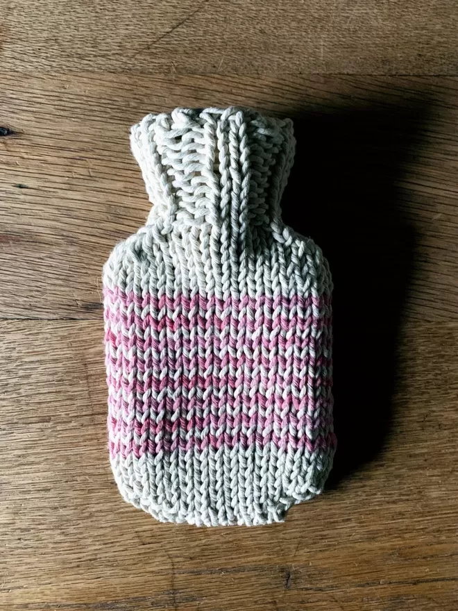 Mini Hot Water Bottle with pink striped knitted string cover on an oak table