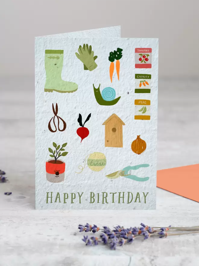 Happy Birthday Seeded Card with Gardening Illustrations including wellies, gloves, carrots, bird box, scissors, plant pot, onion, radish, seed packets, a snail and string with a sprig Lavender in the foreground