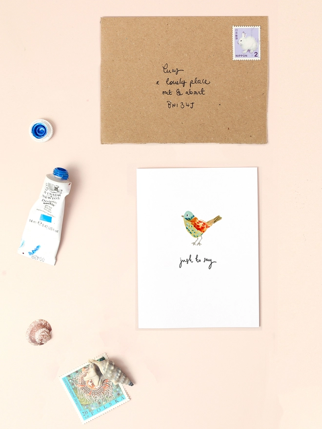 An image of a mini, rectangular white greetings card with an illustration of a colourful bird with a hand written 'just to say' message under the central illustration. The mini greetings card is placed on top of a rectangular brown kraft envelope.