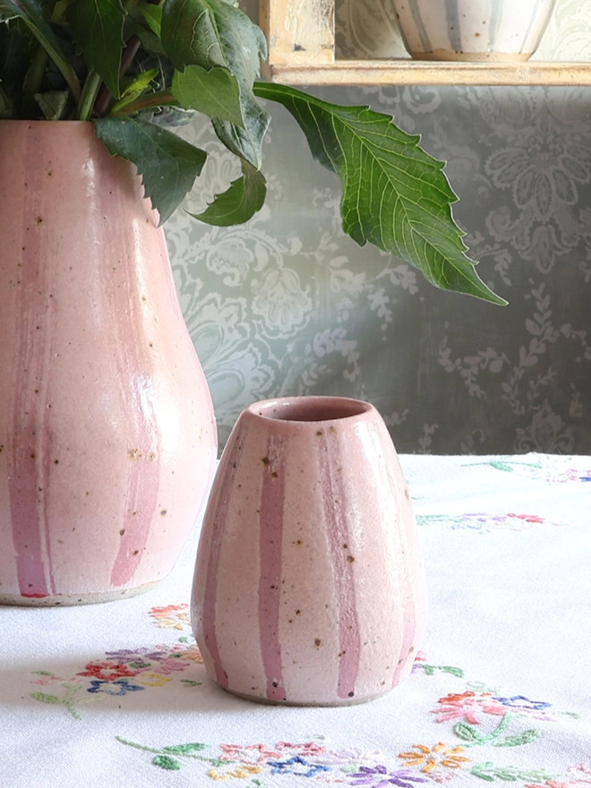 Handmade Ceramic Stoneware Small Vase in Pink on a table with a vintage tablecloth. Behind the vase sits a large vase in the same style. 