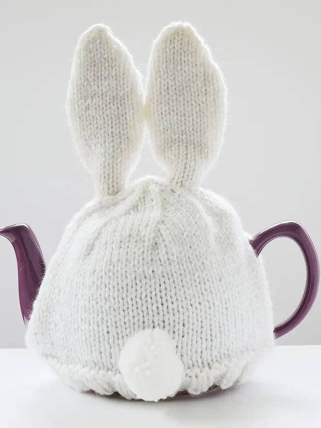 Hand Knitted Bunny Rabbit Tea Cosy, a rabbit bunny tea cosy sitting on a grey surface against a white backdrop. It fits over a purple tea pot. 