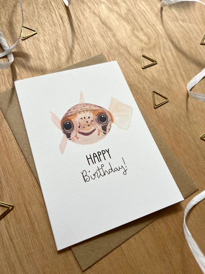 A greetings card with a white background featuring a deflated pufferfish on their back with the phrase "happy birthday" underneath
