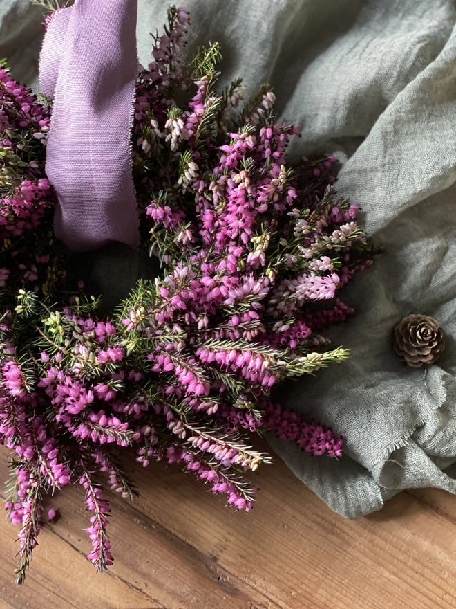 A close up picture of a Petite Wild Heather Wreath in purple and green hues, with a soft lilac ribbon looped through the middle, on display atop a sage green fabric