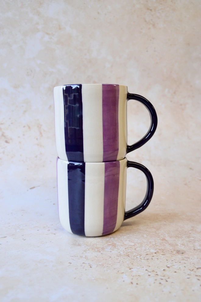 Two handmade striped ceramic mugs stacked on top of each other in purple, cream and dark plum tones, with dark handles, photographed on a neutral stone background.