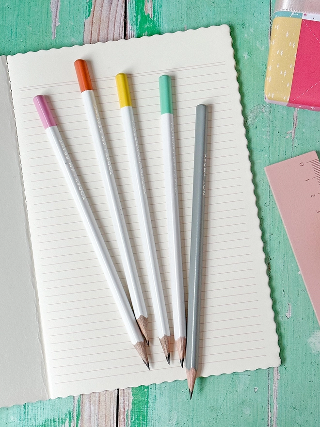 Five pencils with pastel coloured ends lay on an open lined notebook on a green desk.