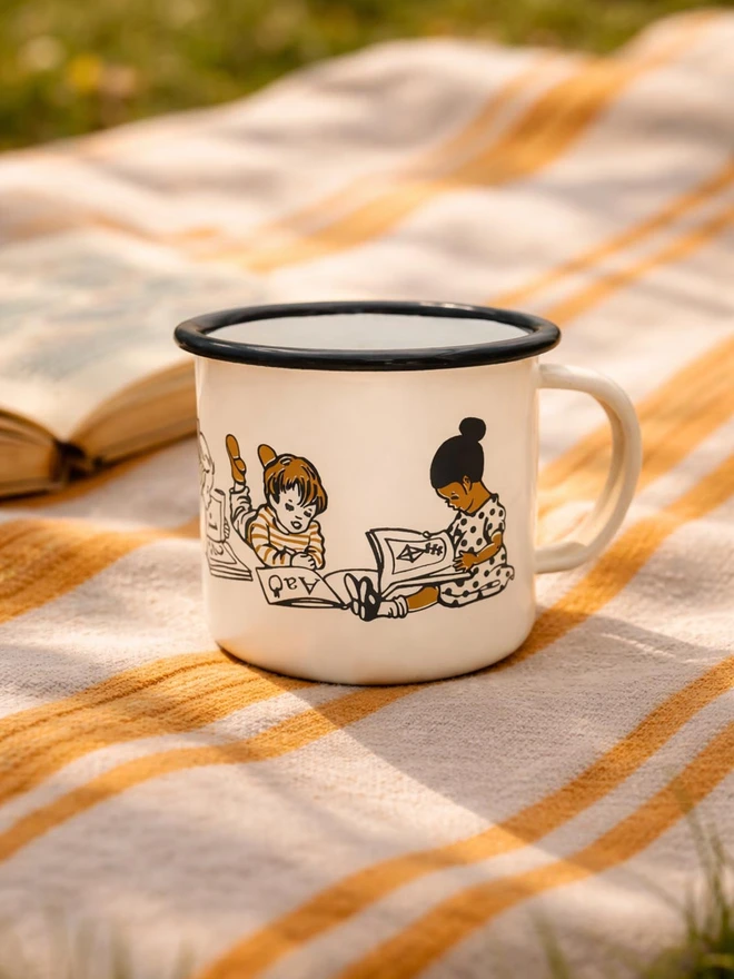 a childrens enamel mug featuring vintage illustrations of children reading placed on a stripped picnic blanket