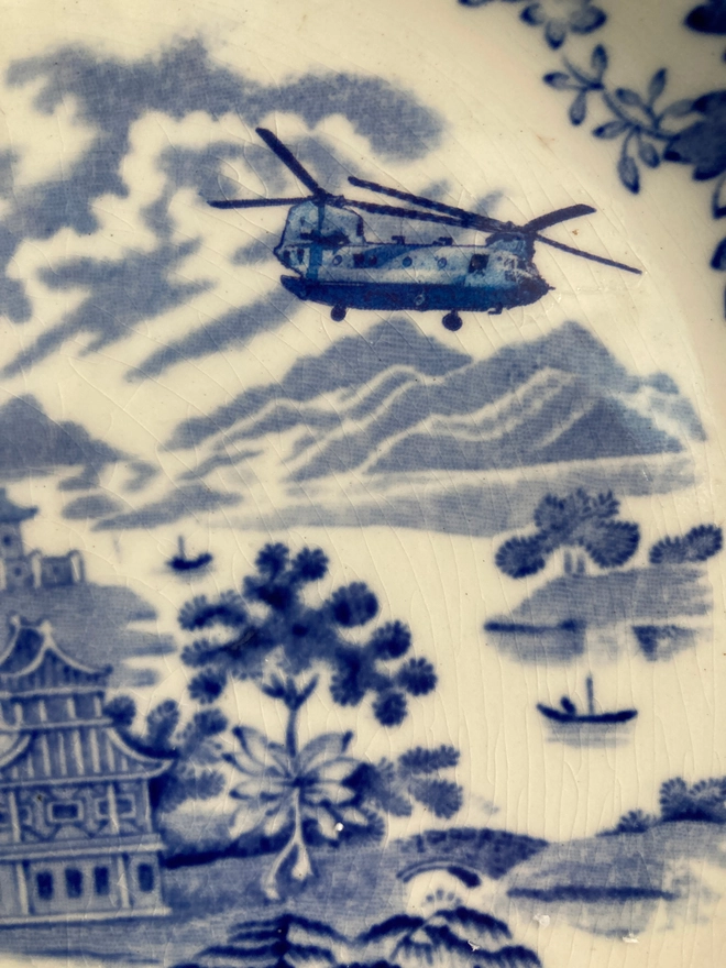 A Chinese style blue and white china plate portrays three people standing near a property. One is holding a fishing rod. A Chinook helicopter flies overhead.