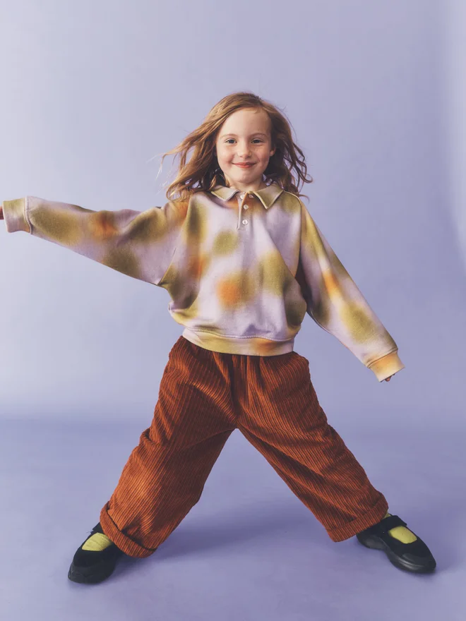 Child stands with arms outstretched against a lilac backdrop, wearing the Studio Koter Luka jersey top in Hazy Lilac with Ever trousers in umber cord.  Two earlier ones can be updated too: 0402 and 0426 are also Luka in Hazy Lilac with Ever trousers.