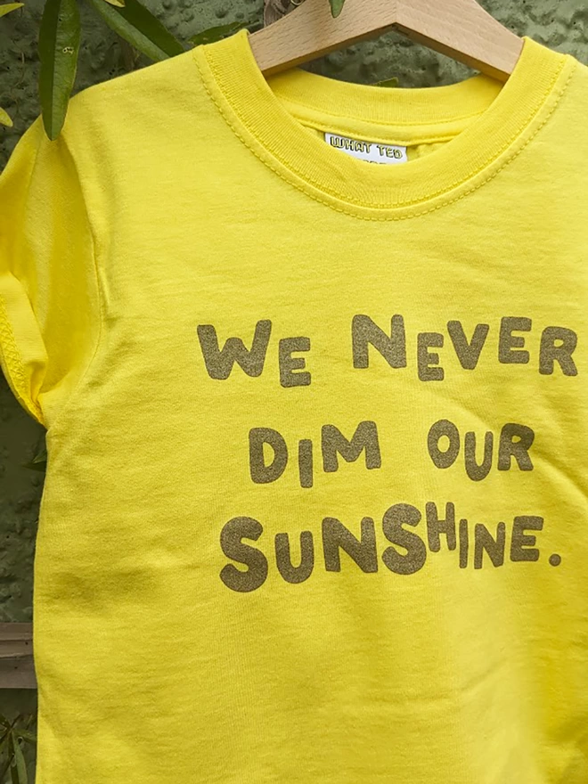 A yellow t-shirt with the slogan We Never Dim Our Sunshine hanging on a garden trelis