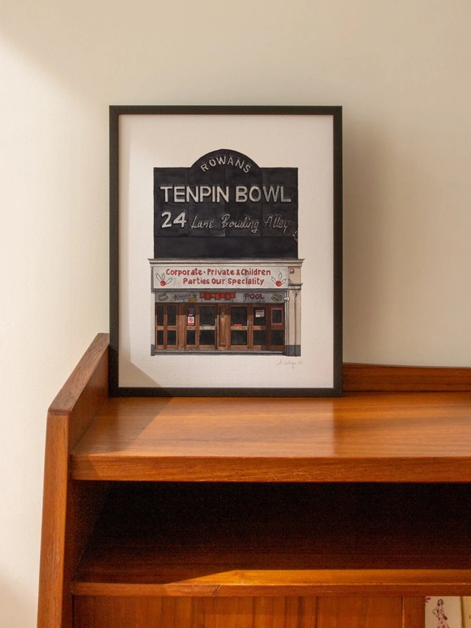 A framed watercolor print of Rowans Tenpin Bowl displayed on a mid-century wooden cabinet.