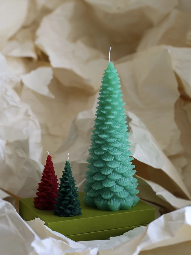 Large Christmas Tree Candle. a large mint green christmas tree shaped candle stands on a green box, nestled among crumpled cream tissue paper. a smaller red and a smaller darker green tree candle sit in front of it for scale, both with visible wicks. the textured, layered pine branch design and slightly variegated wax give all three candles a realistic, hand-poured finish.