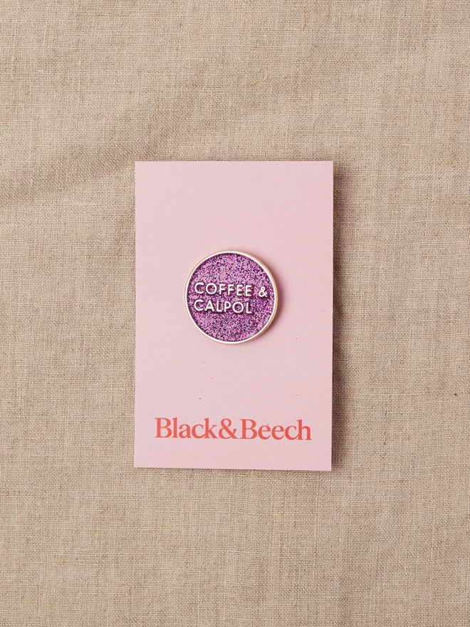 A round purple glitter enamel pin with the words Coffee and Calpol written in gold, on pink backing card