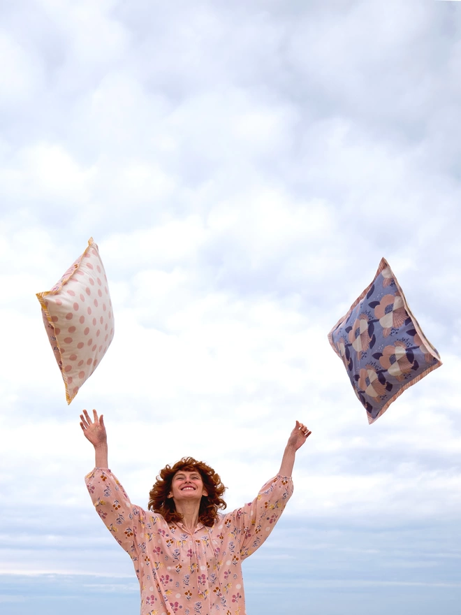 Young woman in dusty pink block printed pyjama throwing two cushions into the air, one featuring  a large scale blue block printed floral and one in a pink spot over a cream background 