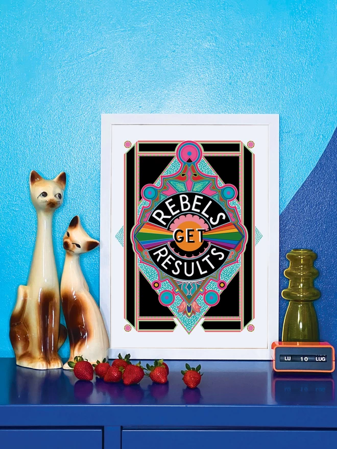 Rebels Get Results is written in white on a black background at the centre of this vibrant, abstract portrait illustration, with a black background and rainbows emitting from the centre , and blue and pink detailing. The picture is in a white frame, against a turquoise and blue wall resting on a blue cabinet. Next to the picture are two cat ornaments, some ripe strawberries, a yellow glass vase and an orange Italian plastic calendar showing the date as ‘LU 10 LUG’.