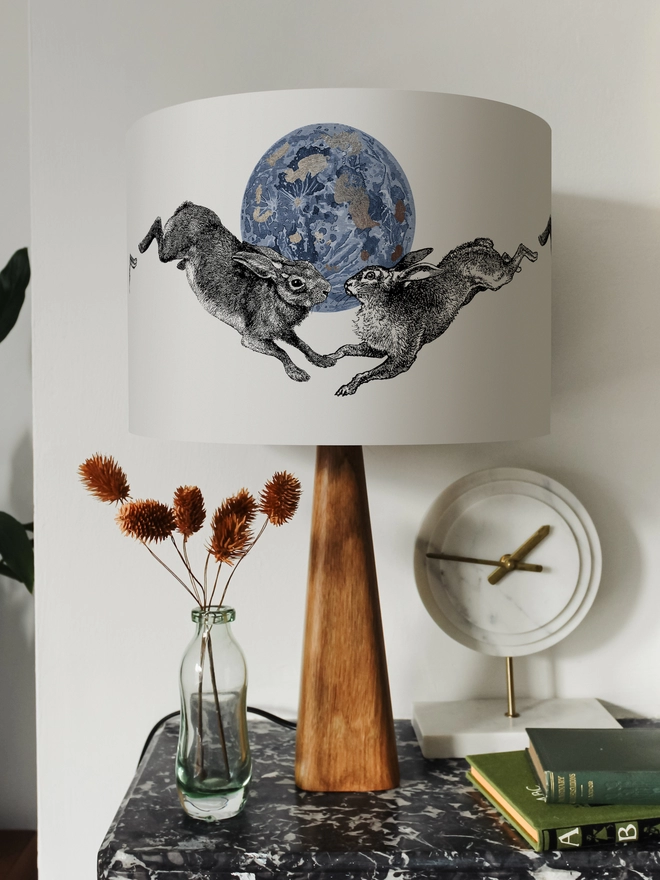 Drum Lampshade featuring a pair of hares leaping across a blue and silver moon on a wooden base on a shelf with books and ornaments