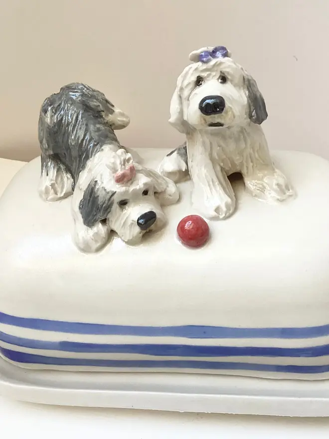 Two white and black dogs with bows in their hair on a butter dish. One is lying down, one is sitting. The base is white with three blue stripes around the bottom.