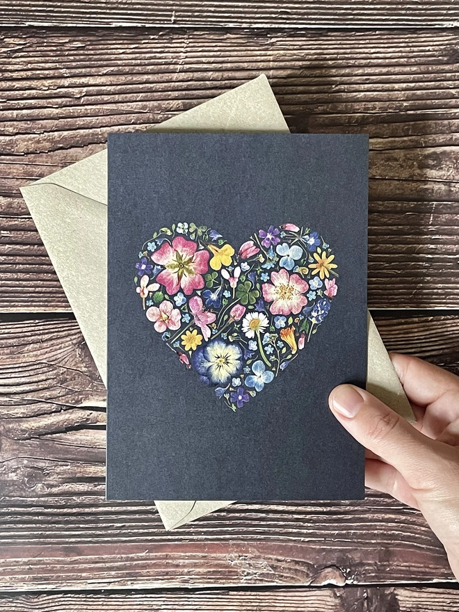 Hand Holding Greetings Card with Digitally Printed Pressed Flower Heart Design - Brown Kraft Envelope - Brown Wooden Background