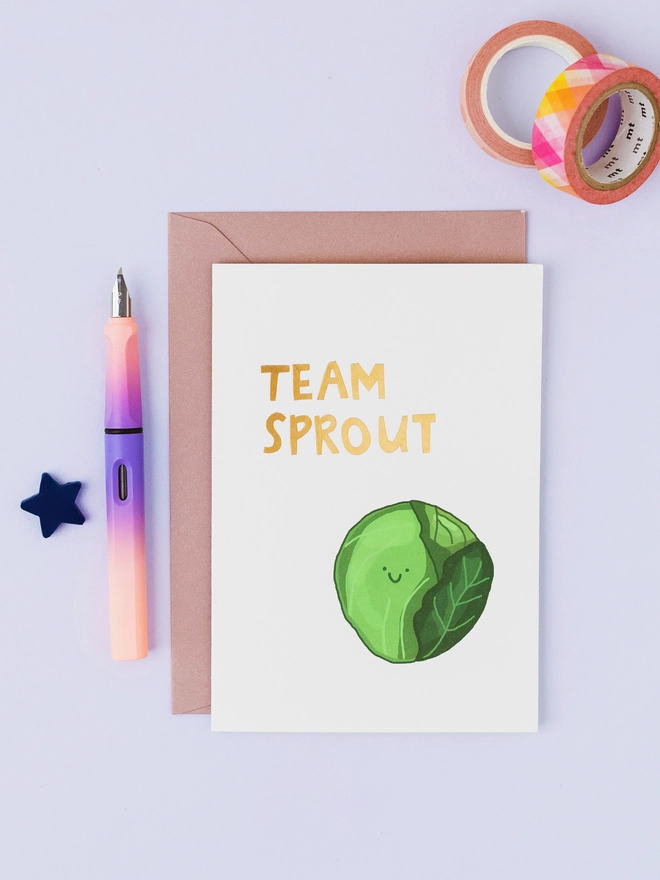 White Christmas greeting card featuring a hand-drawn green Brussels sprout with a smiling face and gold foil Team Sprout lettering.