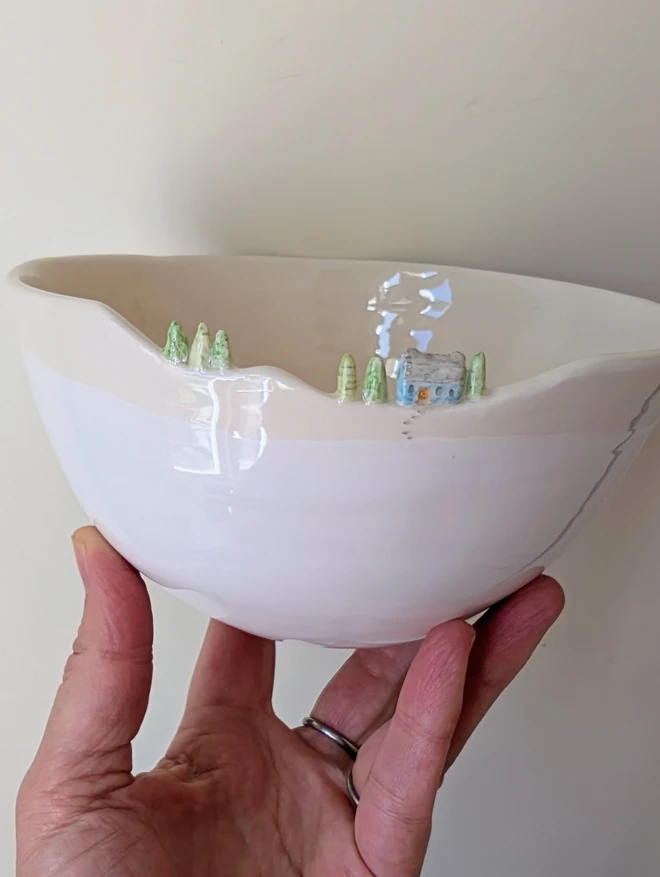 A handmade ceramic white bowl with a tiny blue house and tree details on the rim and white glaze 