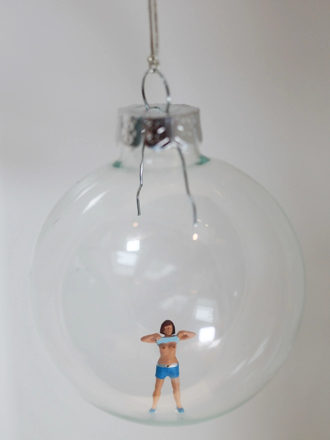 Miniature figure of woman playfully pulling up her top in a glass Christmas bauble
