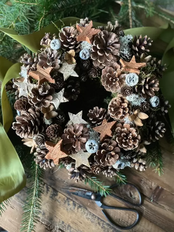 Pine Cone Wreath decorated with natural bark stars, sits on a wooden table, green foliage and a lighter thick green ribbon surrounds the wreath.