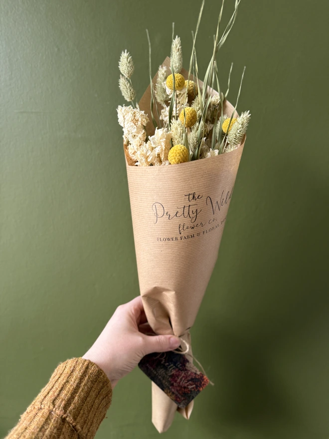a bunch of dried flowers in bright colours wrapped in black tissue and kraft paper held by a person in a mustard coloured jumper with their left arm against a green wall