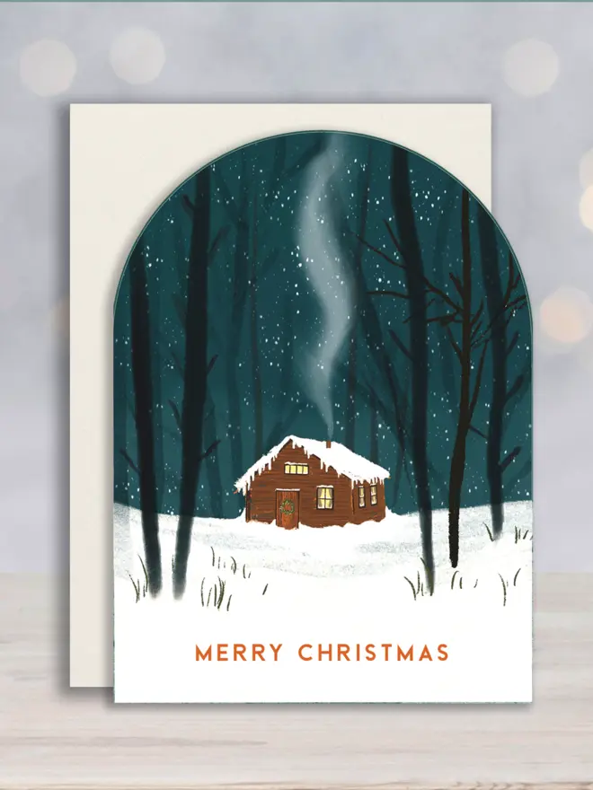 Arch shaped Christmas card depicting an illustrated Cabin in the snowy woods, message reads Merry Christmas.