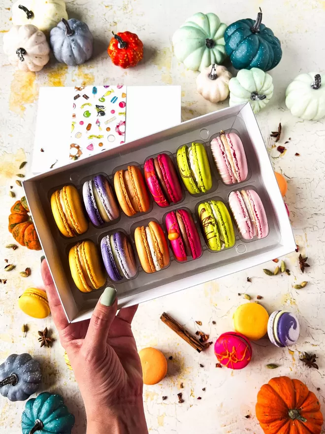 a person holding a box of colourful autumn macarons surrounded by colourful pumpkins on a table 