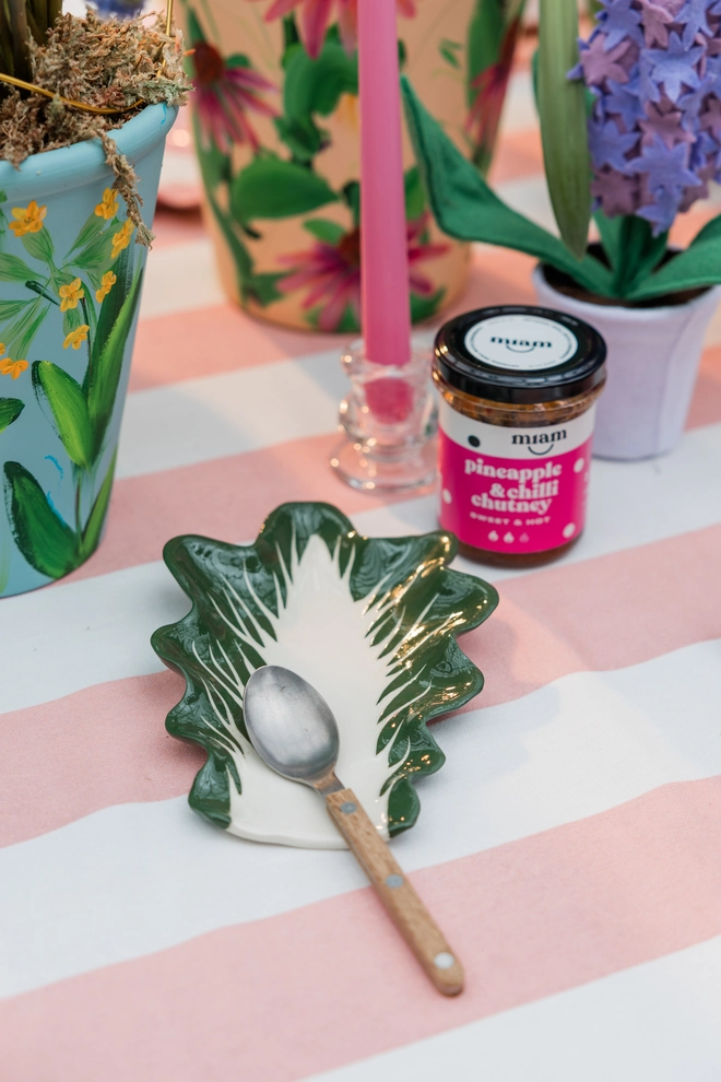Radicchio Leaf Spoon Rest in Dark Green as part of a larger tablescape with a pink and white tablecloth, painted plant pots and colourful cutlery