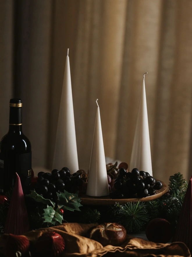 White Christmas Tree Cone Candles (Set Of 3). Three unlit cream cone candles on a wooden board with black grapes, pine garland, holly and a bottle of red wine. The tapered candles add height and understated elegance to a Christmas dinner table.