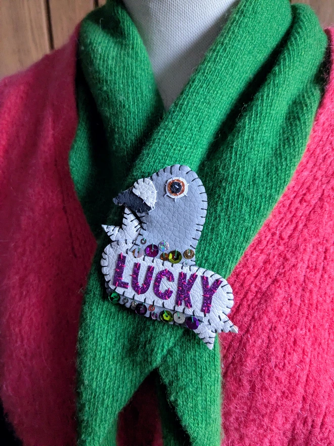 Hand-stitched white and grey faux leather pigeon brooch with purple glitter ‘LUCKY’ lettering and colourful sequin details, pinned to a bright green scarf