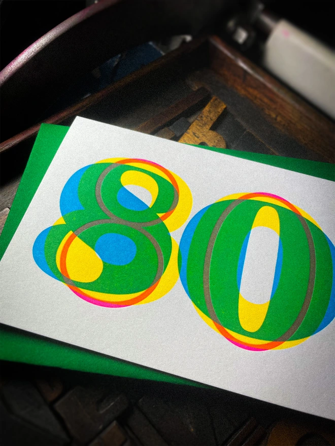 Handmade letterpress 80th birthday card featuring bold overlapping numerals printed in vibrant fluorescent cyan, magenta and yellow inks. Each card is individually hand printed using traditional letterpress techniques on luxury paper stock, creating a beautifully tactile finish with a subtle deboss and rich layered colours inspired by the CMYK printing process.