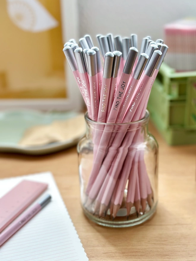 A jar of pink pencils stands on a wooden desk surrounded by stationery items. Each pencil has the words Find The Joy on the side.
