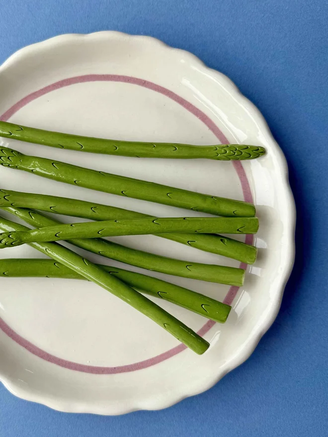 Ceramic white plate with pink trim, topped with ceramic fake asparagus tips on a blue background