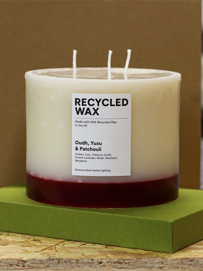 Oudh, Yuzu & Patchouli Candle (Set Of 1 Or 3 Wicks). the oudh, yuzu and patchouli candle stands on a green box against a brown background, showing its three wicks and cream wax with a deep red base. a white label reads "recycled wax, made with 50% recycled wax in the uk", followed by the scent name and note list of amber, yuzu, tobacco, oudh, french lavender, musk, patchouli and bergamot. the candle's two-tone colouring and visible drip texture give it a rustic, hand-poured look.
