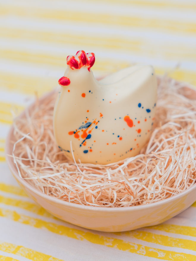 A white chocolate chicken from Nenette Chocolates, sitting in some straw in a yellow bowl on a yellow and white striped tablecloth