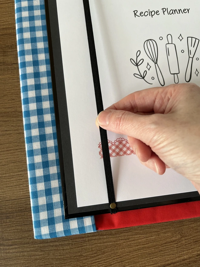 Close-up of hand holding black elastic riveted inside ring binder, that holds recipe planner page in place. Blue gingham and red lined fabric binder cover lies beneath.