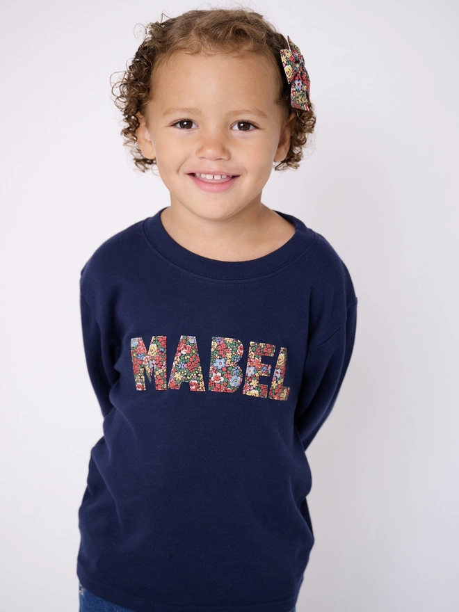 a 3 year old girl wearing a navy long sleeve t-shirt with her name Mabel cut from Liberty floral fabric and sewn onto the front 