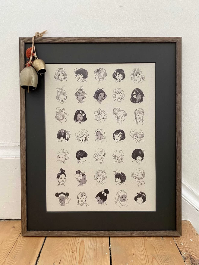 A3 poster print of vintage girls wearing different styles of hairbows shown in dark oak frame with black mount propped on floor with brass bells
