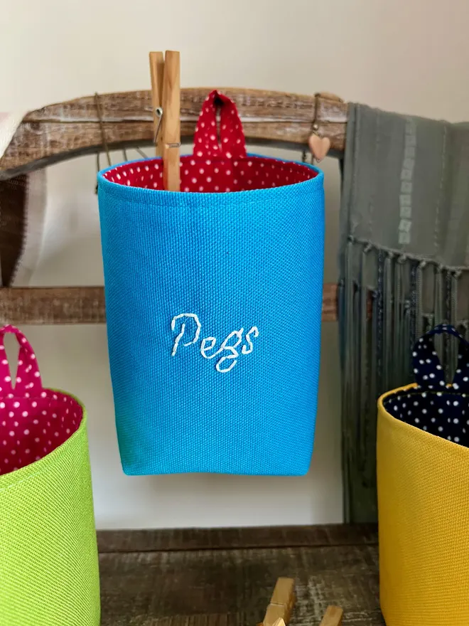 Blue storage bag hanging on a wooden stand, the front embroidered with ‘Pegs’ in white thread