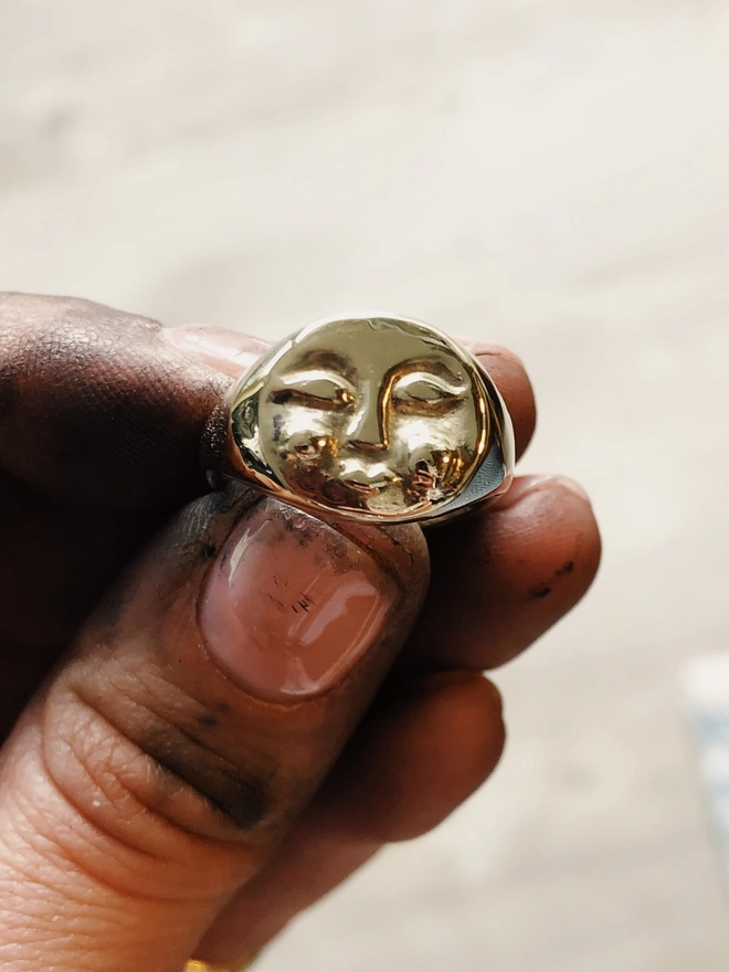 Image of hand carved gold moon face ring being held up by the finger tips of a hand 