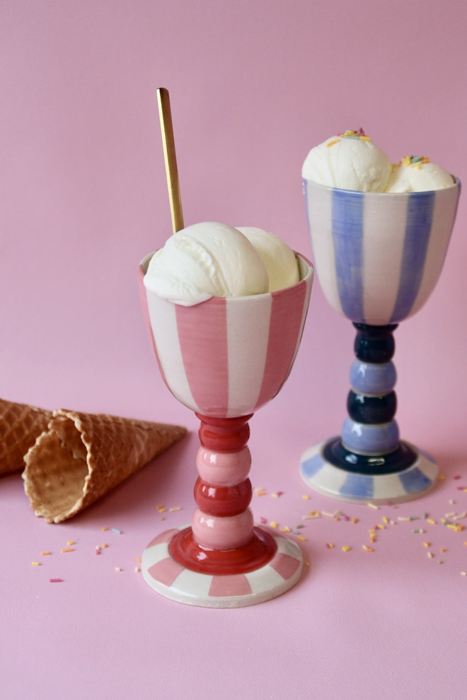 Handmade striped ceramic goblets in pink and blue colourways, filled with scoops of vanilla ice cream and sprinkles, styled on a pink background with waffle cones.