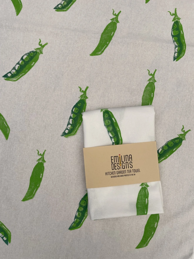 Pea Kitchen Tea Towel. White fabric with a diagonal pattern of green pea pods, some open to show bright green peas.