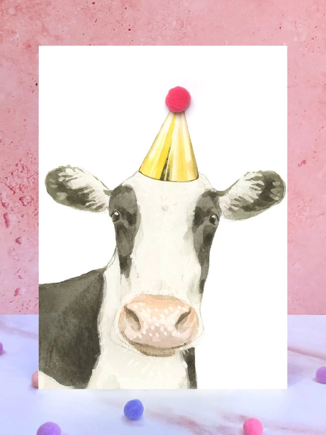 A greeting card featuring a hand painted design of a cow, stood upright on a marble surface surrounded by pompoms. 