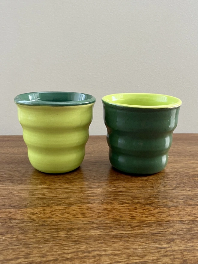 a pair of ceramic tumblers with a wavy sides on a wooden table. the pair is glazed in reverse colours. one is lime green on the outside and forest green on the inside and the other is the opposite with the forest green on the outside and lime green on the inside.  